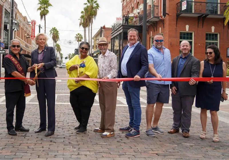 Eight people holding a red ribbon during a ribbon cutting on a brick paved street