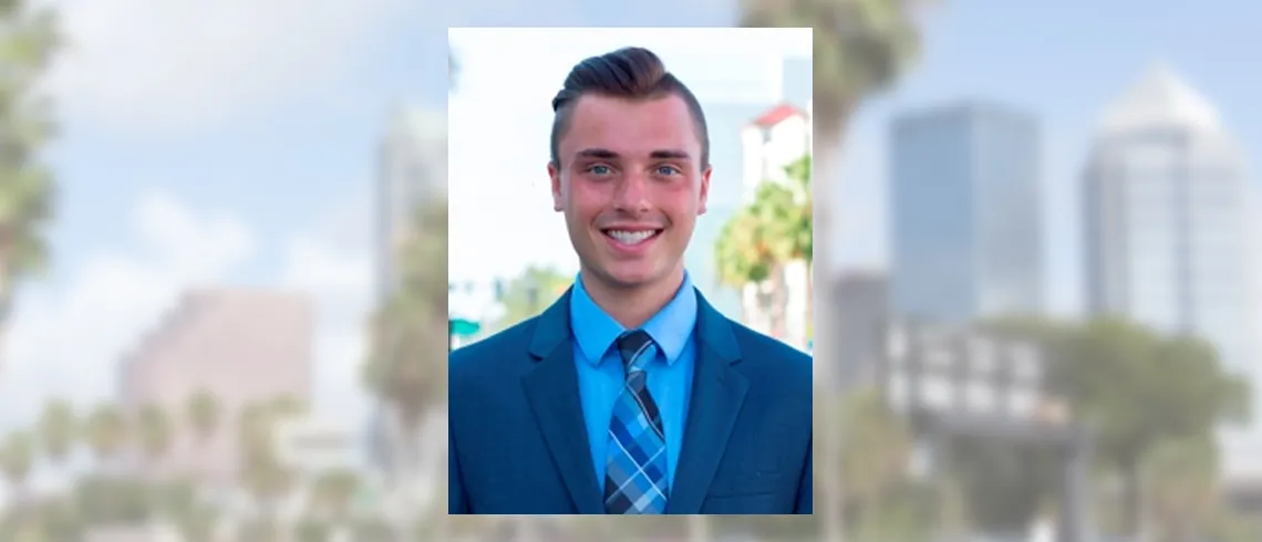 Smiling young man in a blue suit and tie with the Tampa skyline blurred behind him