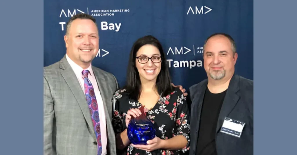 Three people with a blue glass award in front of an American Marketing Association Tampa Bay backdrop