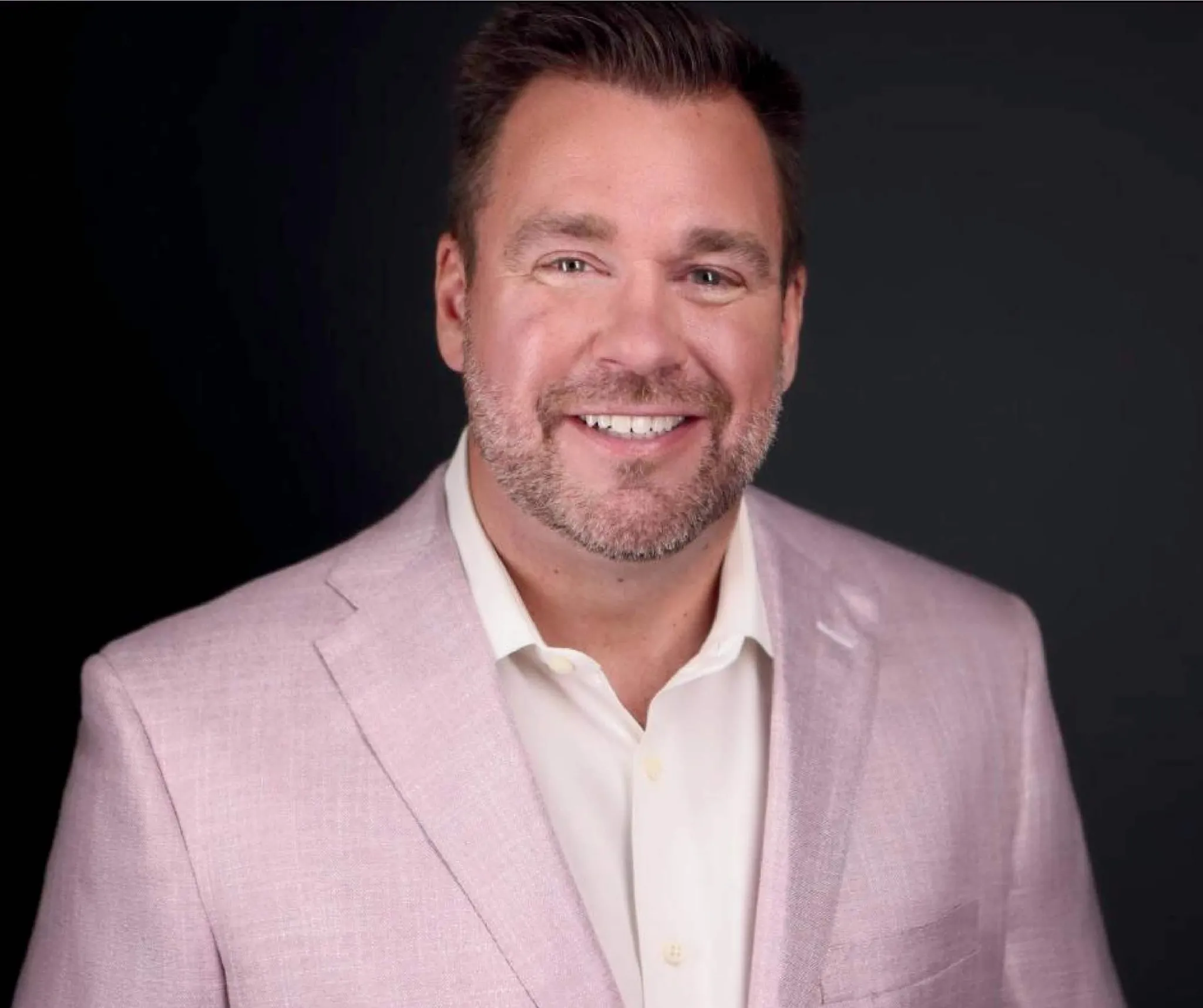 Smiling man with short brown hair and a beard in a pink blazer against a black background