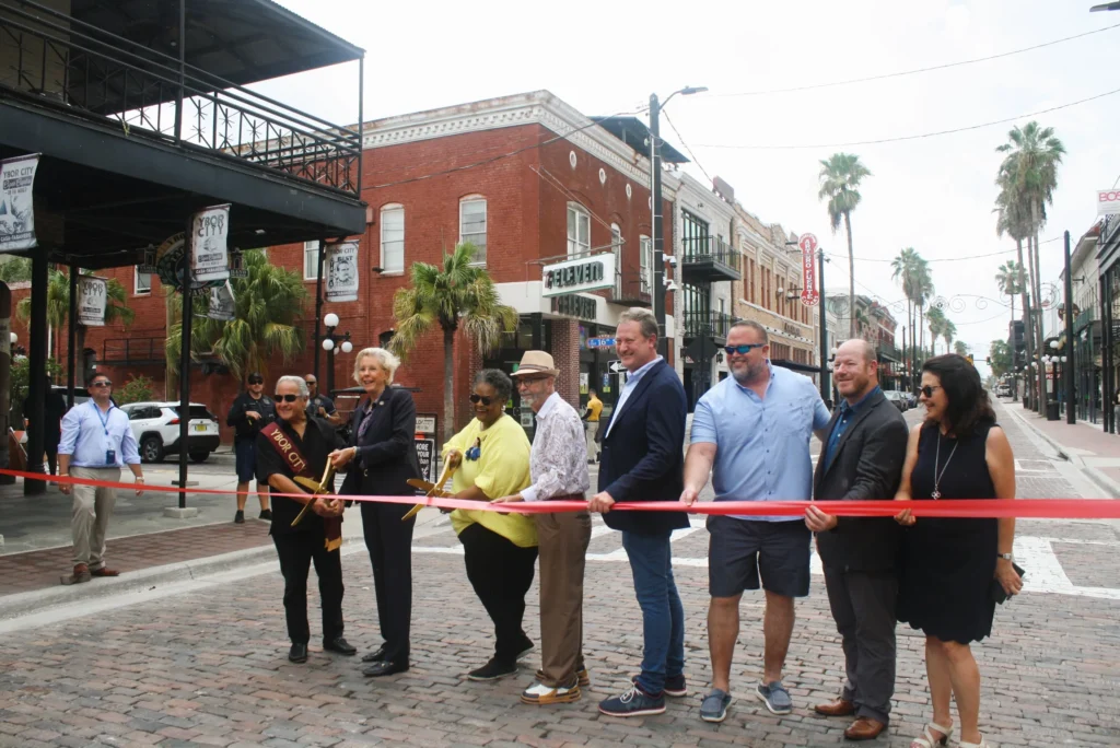 Group of people holding a red ribbon at a ribbon cutting on a brick street in Ybor City