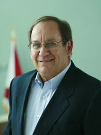 Smiling older man with glasses in a dark blazer and light blue shirt in front of a flag