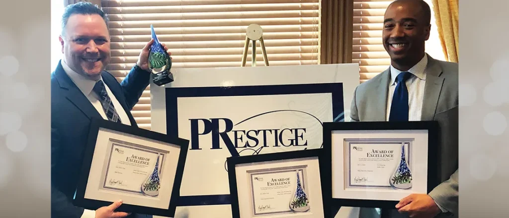 Two men holding framed award certificates in front of a PRestige sign