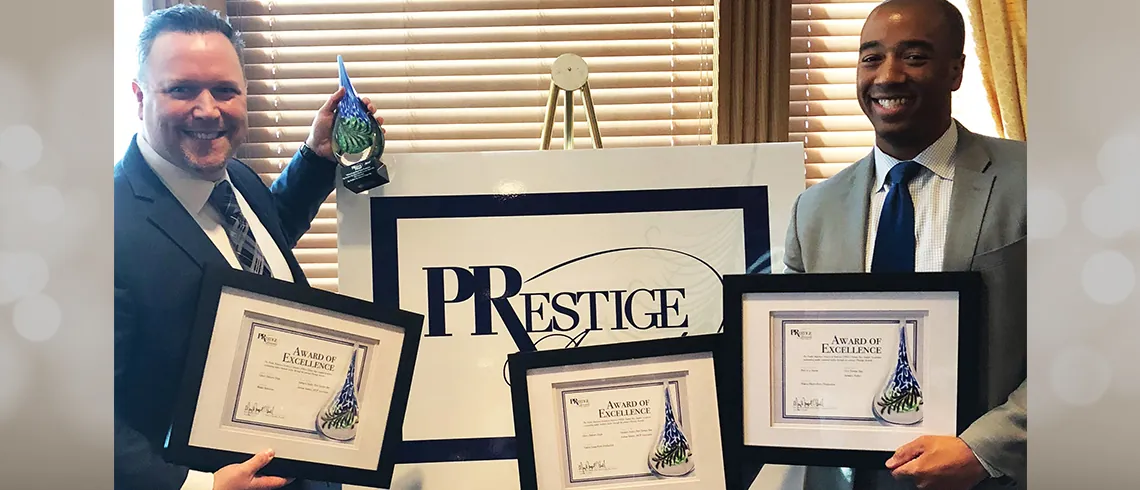 Two men holding framed award certificates in front of a PRestige sign