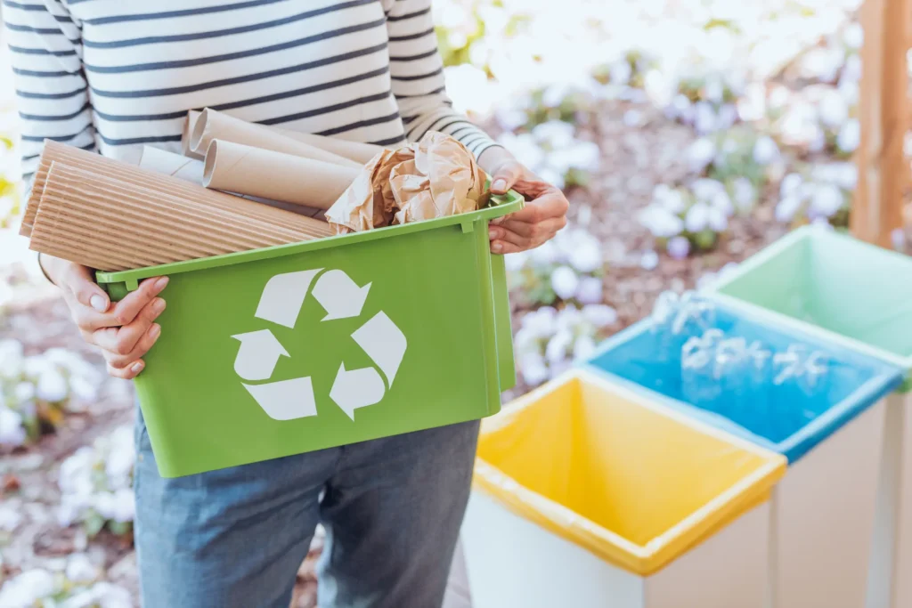 Person holding a green recycling bin with cardboard and paper beside three colored bins