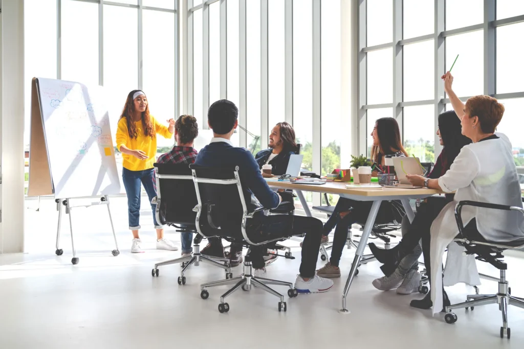 Team meeting around a long table in a bright room with a woman presenting at a whiteboard