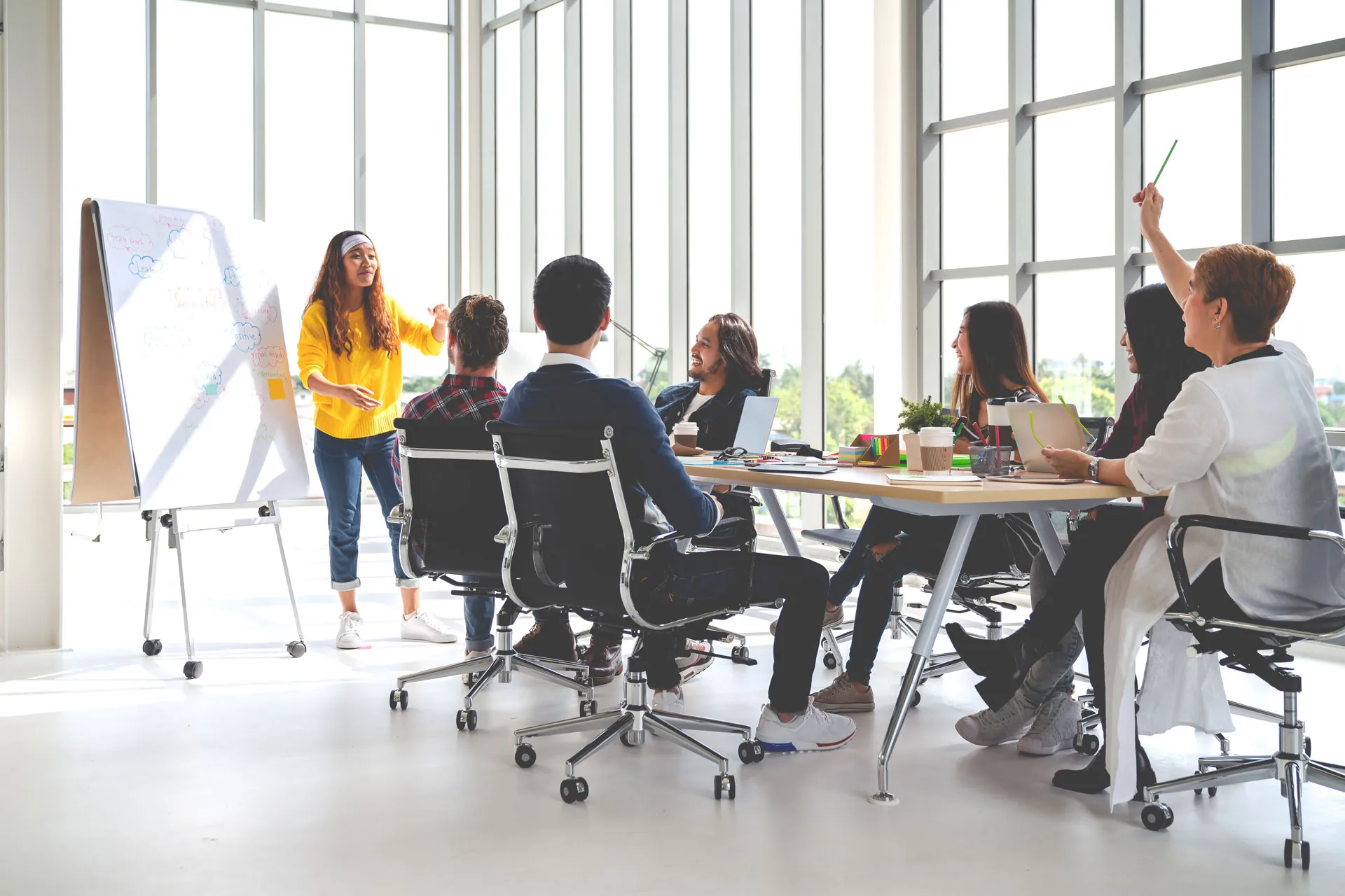 Team meeting around a long table in a bright room with a woman presenting at a whiteboard