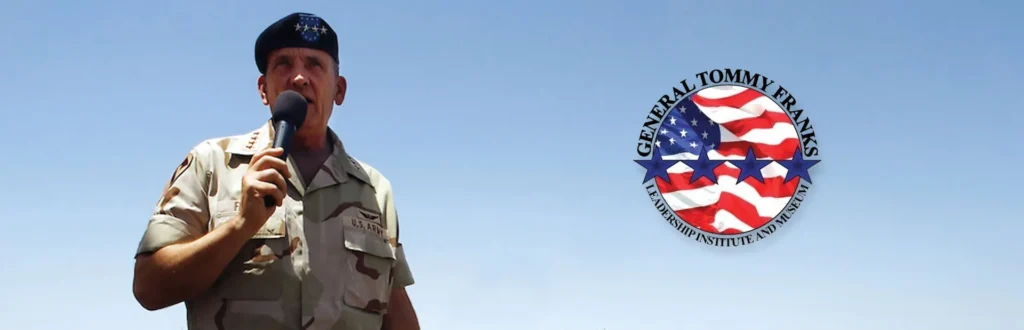 Man in a desert uniform speaking into a microphone, with a General Tommy Franks round emblem