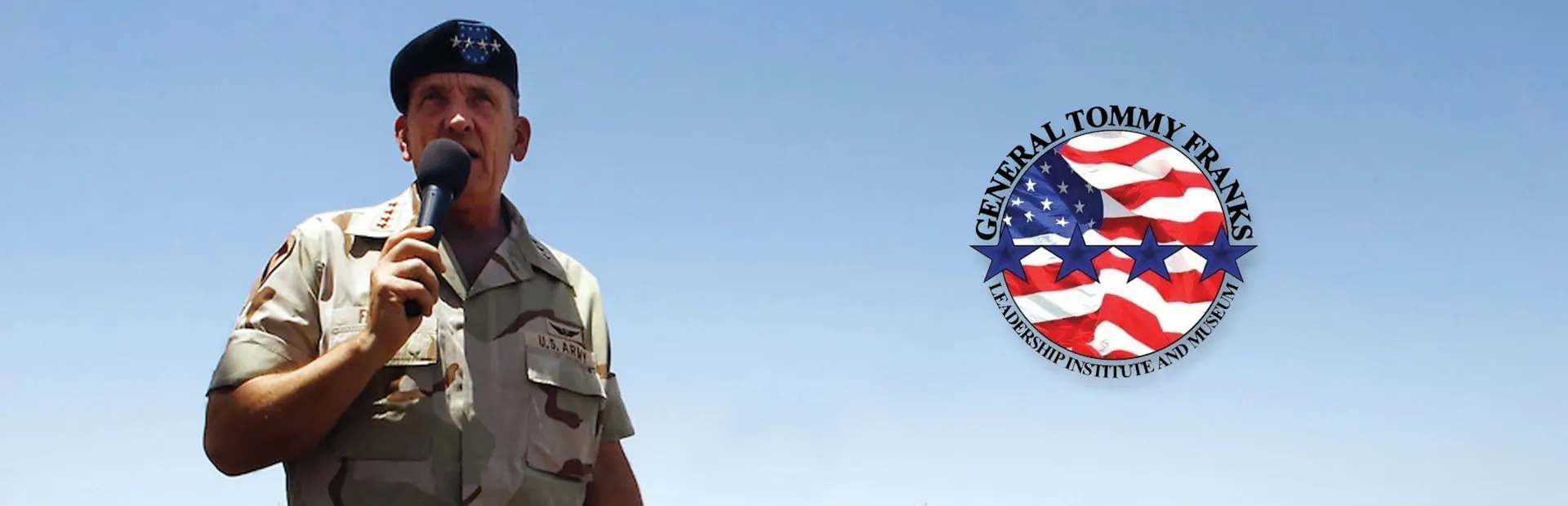 Man in a desert uniform speaking into a microphone, with a General Tommy Franks round emblem