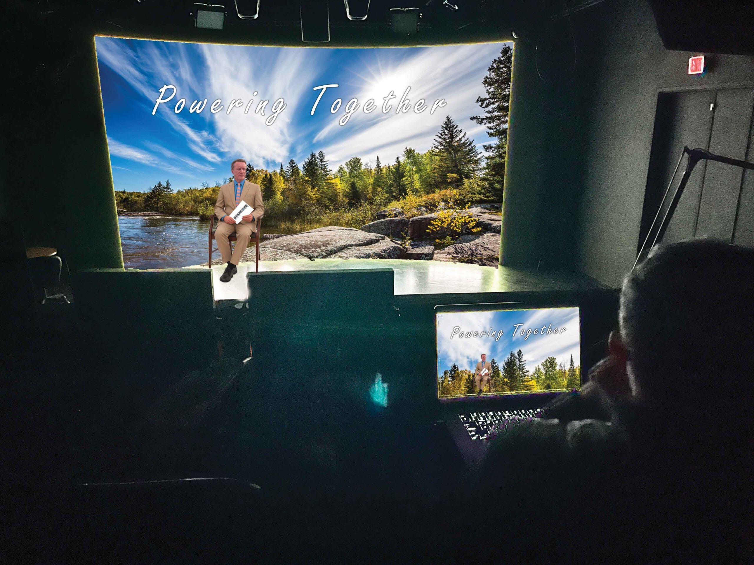 Man seated in a dark studio in front of a large screen showing a lake and forest scene