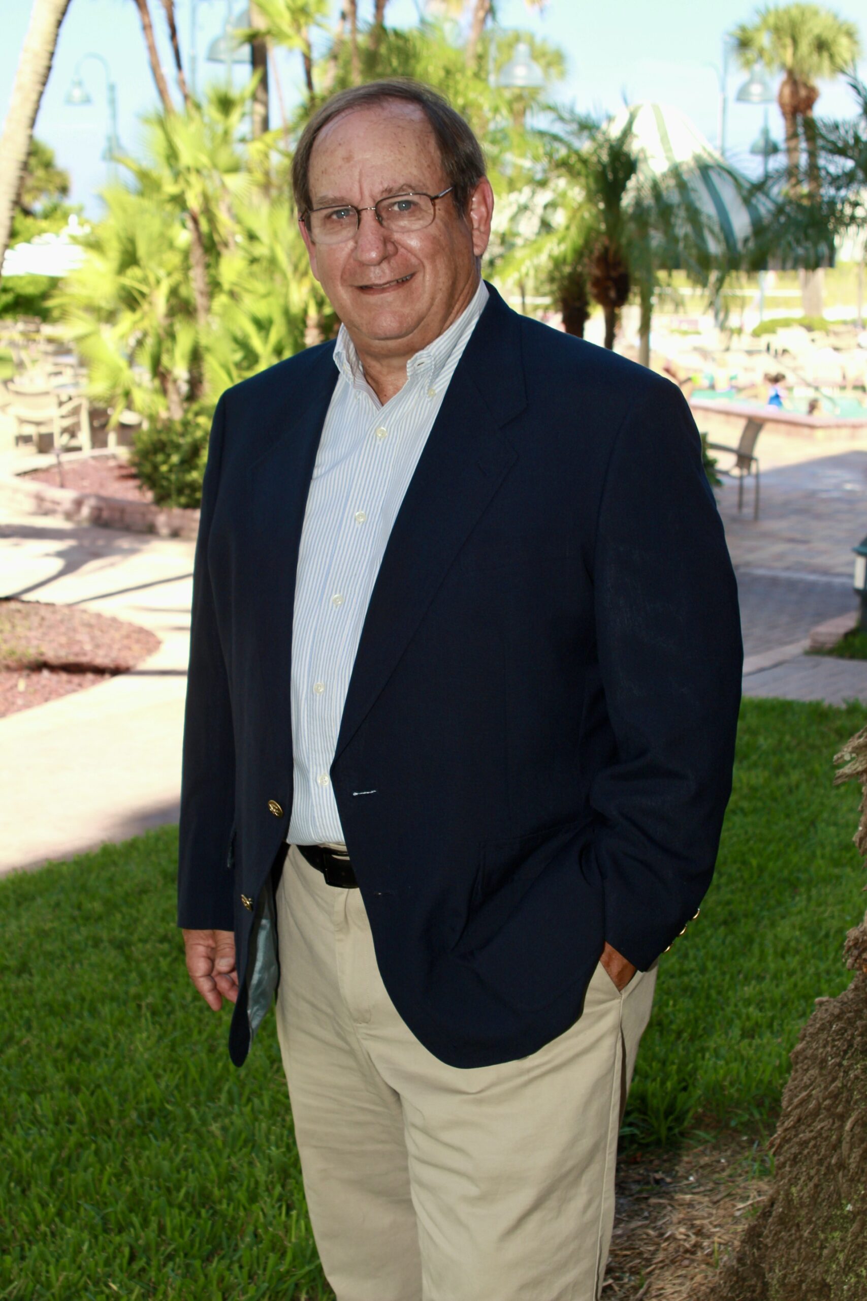Smiling older man with glasses in a navy blazer and khaki pants standing on a lawn with palm trees