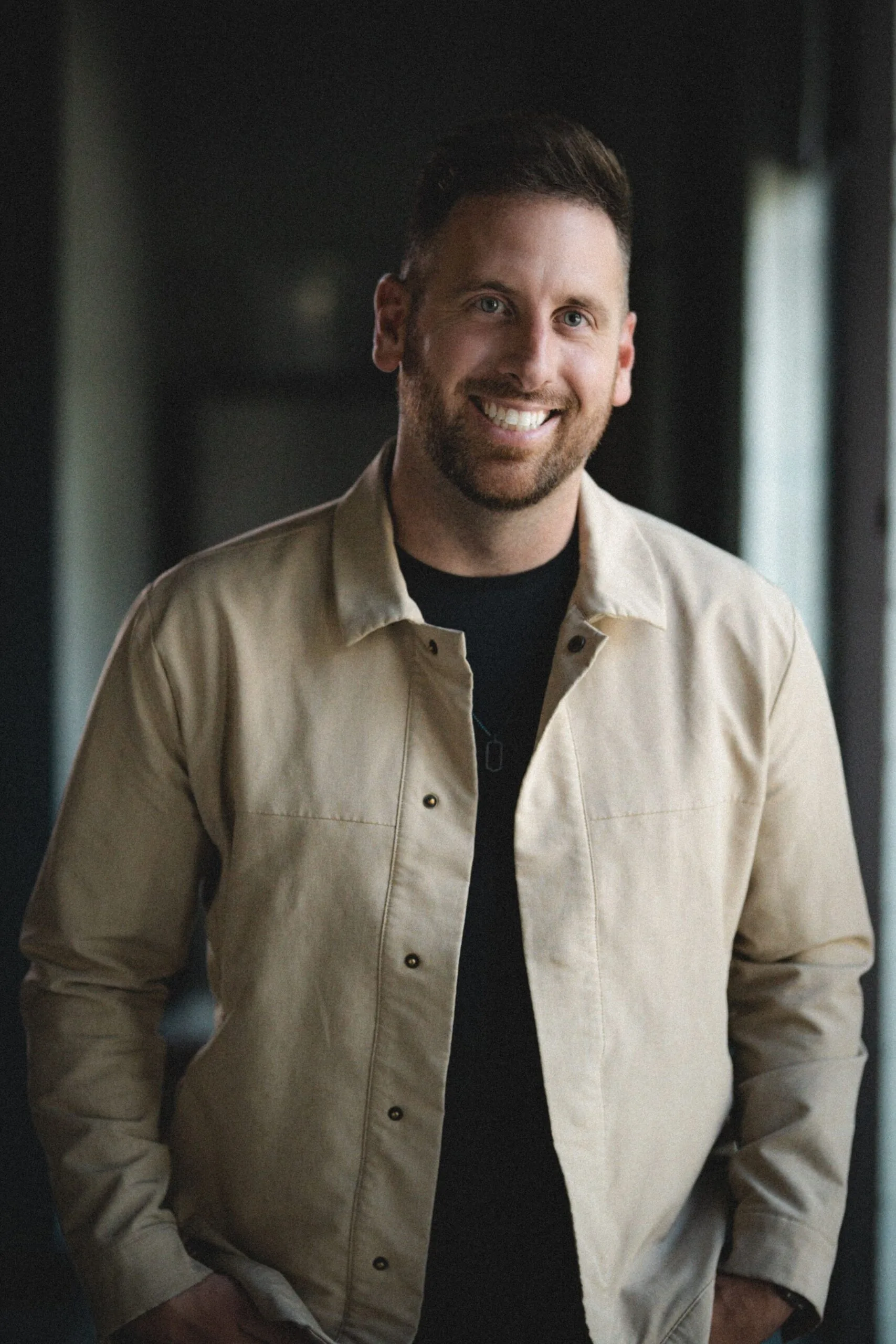 Smiling man with short brown hair and beard in a beige jacket over a black shirt