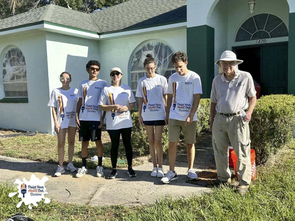 Volunteers posing in front of a white house with an older man in a hat