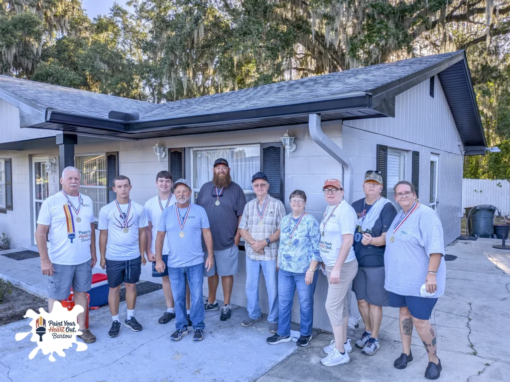 Large group of volunteers posing in front of a gray house with a dark roof
