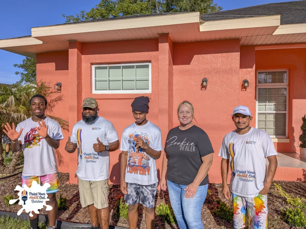 Five volunteers in matching shirts holding paint tools in front of a salmon colored house