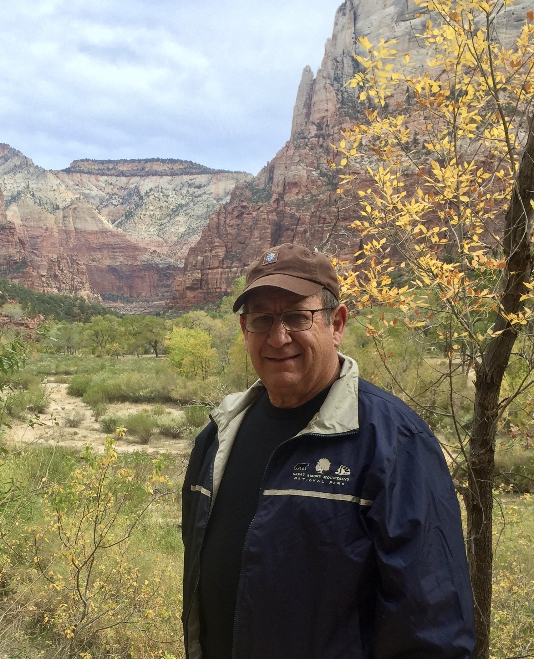 Man in a baseball cap and navy jacket standing among red rock cliffs and autumn trees