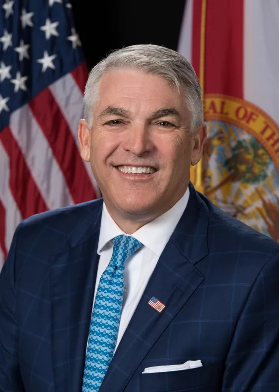 Smiling man with gray hair in a navy suit and teal tie in front of American and Florida flags