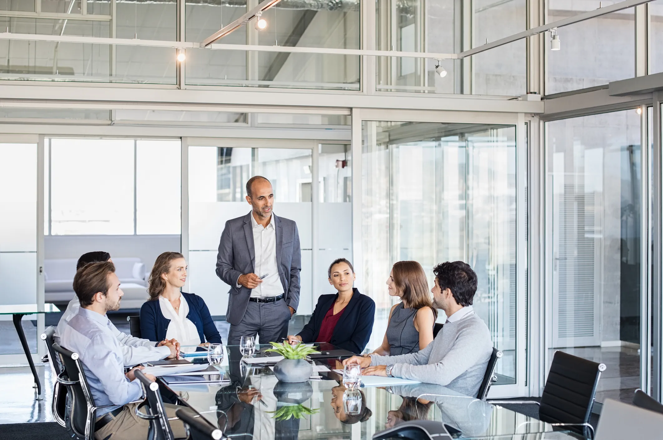 Man standing to address colleagues seated around a table in a glass walled meeting room
