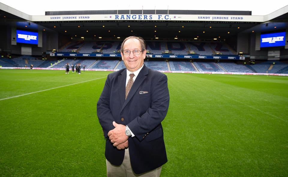 Man in a dark blazer standing on the pitch of a Rangers F.C. stadium