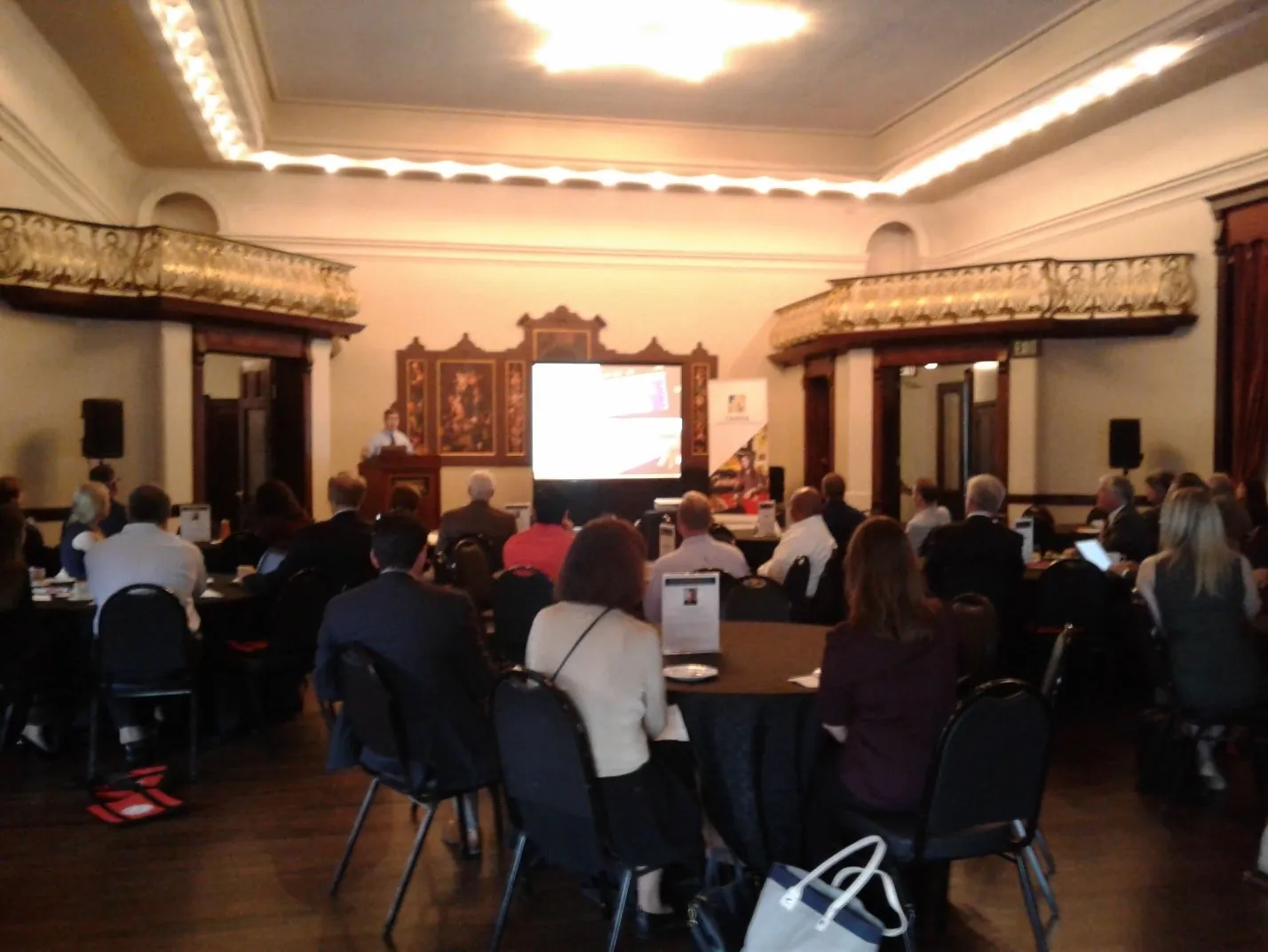 Attendees seated at round tables in a historic ballroom facing a projection screen
