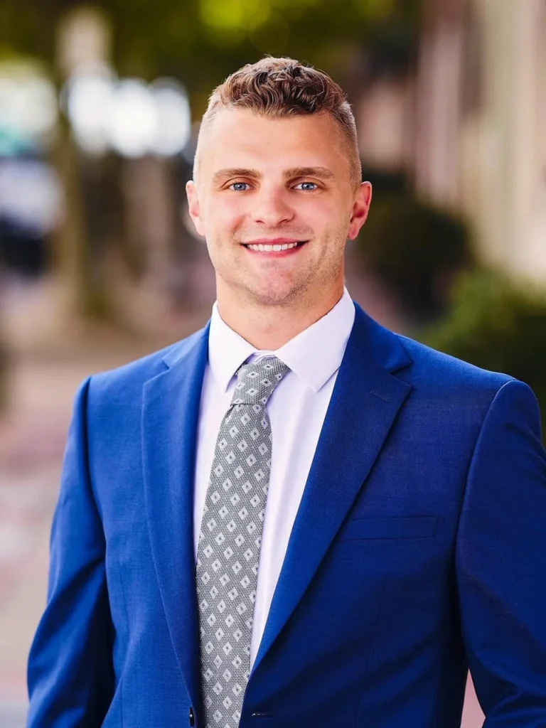 Smiling young man with short light brown hair in a blue suit and gray patterned tie