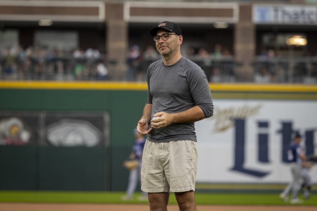 Man in gray shirt and khaki shorts standing on a baseball field before a game