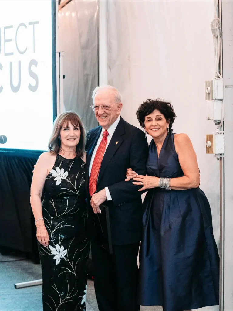 Man in a dark suit standing between two women in evening dresses in front of a Project Focus backdrop