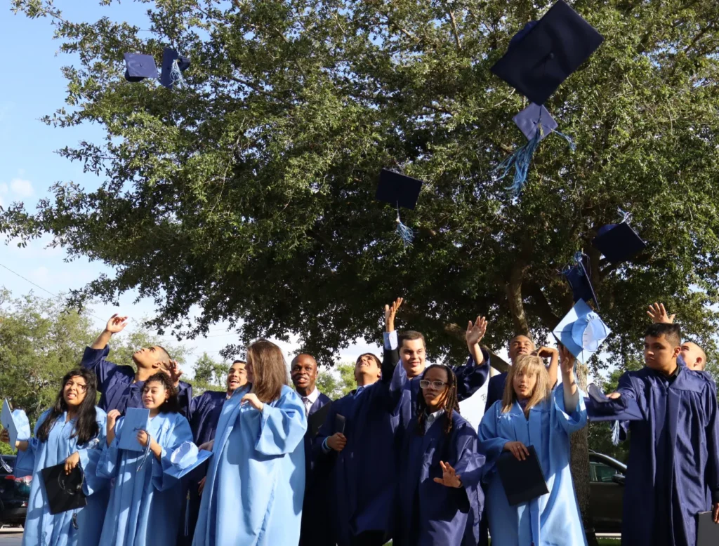 Graduates in blue gowns tossing their caps in the air under a tree