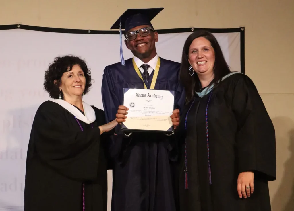 Graduate in cap and gown holding a certificate between two women in black gowns