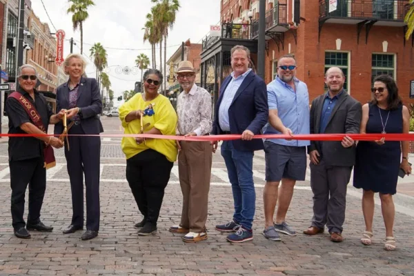 Eight people holding a red ribbon during a ribbon cutting on a brick paved street