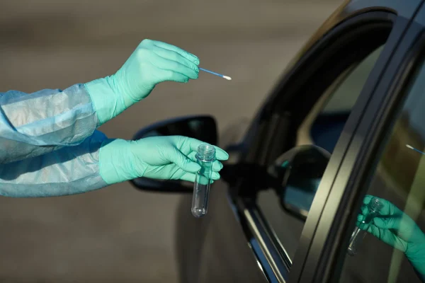 Gloved hands holding a test swab and tube at a car window for drive-through COVID testing