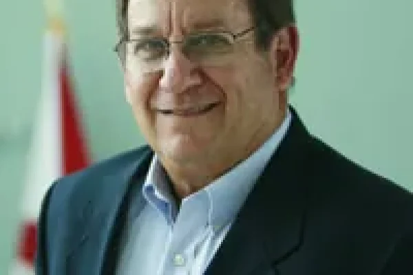 Smiling older man with glasses in a dark blazer and light blue shirt in front of a flag