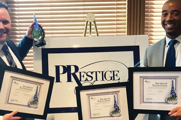 Two men holding framed award certificates in front of a PRestige sign