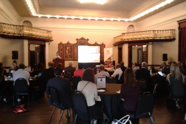 Attendees seated at round tables in a historic ballroom facing a projection screen