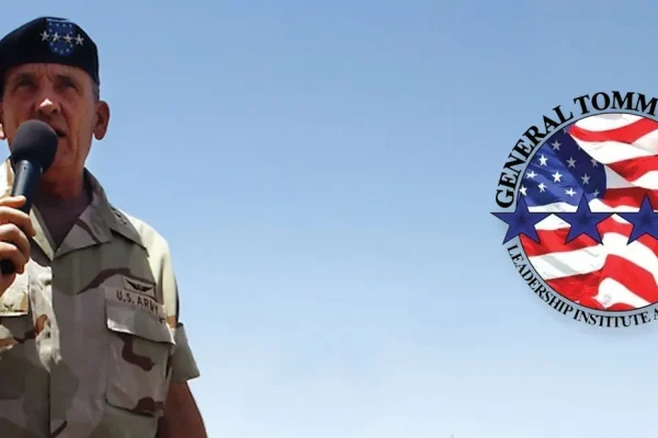 Man in a desert uniform speaking into a microphone, with a General Tommy Franks round emblem